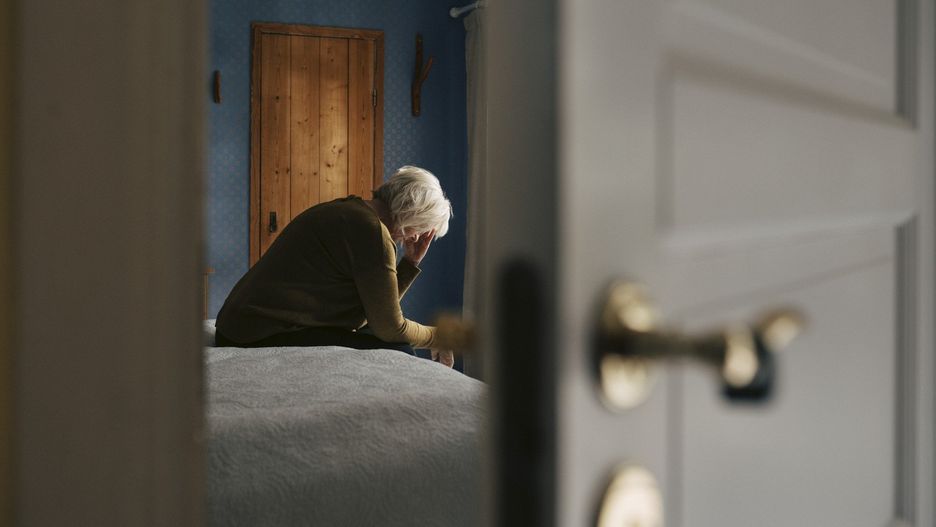 Depressed senior woman sitting with face down on bed seen through bedroom door