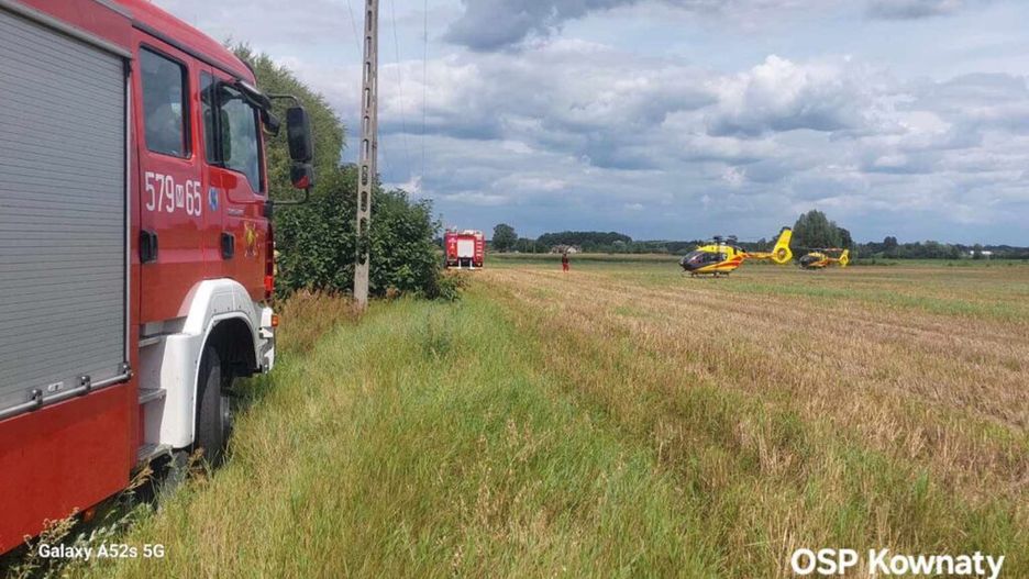 Czerwony wóz strażacki stoi na łące obok dwóch helikopterów ratunkowych, a w tle widoczni ratownicy.