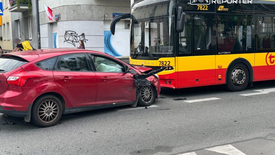 Czerwony samochód osobowy z uszkodzoną przednią częścią zderza się z autobusem na miejskiej ulicy.