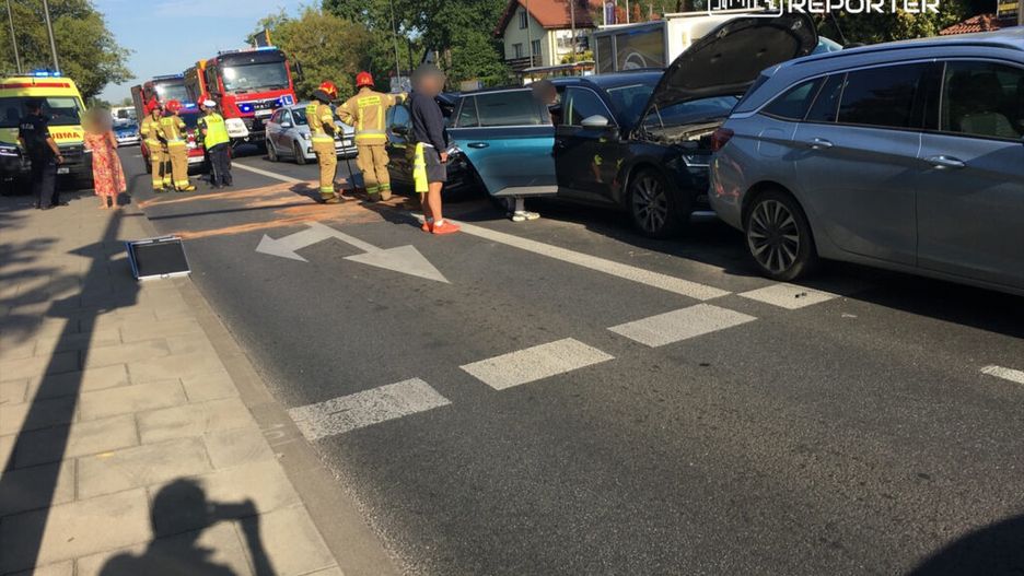 Grupa strażaków i policjantów udziela pomocy na zablokowanej drodze po kolizji dwóch samochodów.