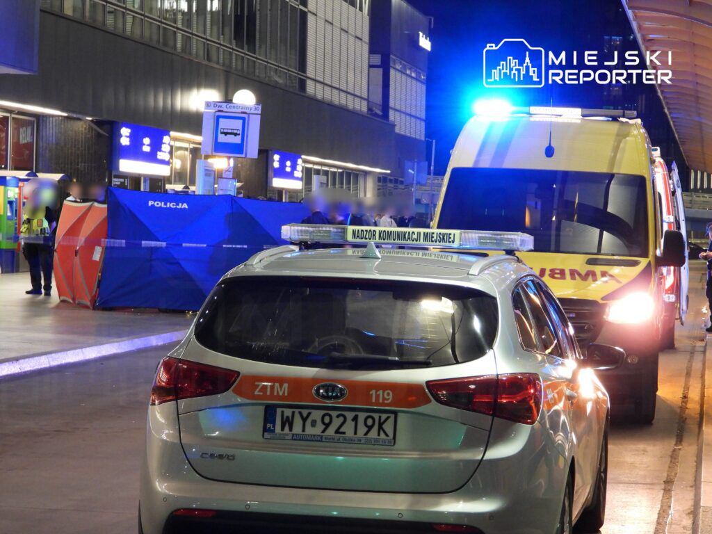 Policyjny namiot zabezpieczający oraz radiowóz z włączonymi sygnałami świetlnymi na stacji kolejowej w nocy.