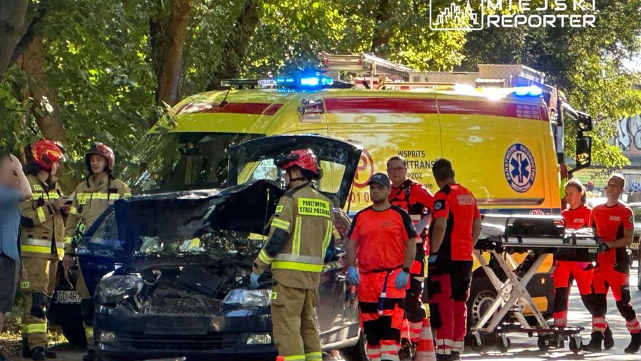 Ratownicy medyczni i strażacy pracują przy uszkodzonym samochodzie na drodze, obok stoi karetka z włączonymi sygnałami.
