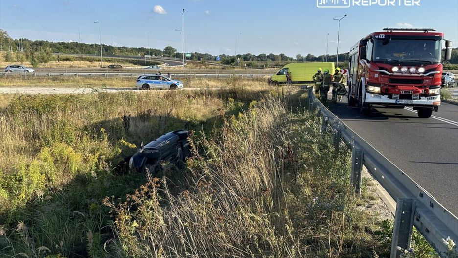 Na poboczu drogi leży przewrócone auto, obok stoją strażacy oraz woz strażacki, a w tle przejeżdżają inne pojazdy.