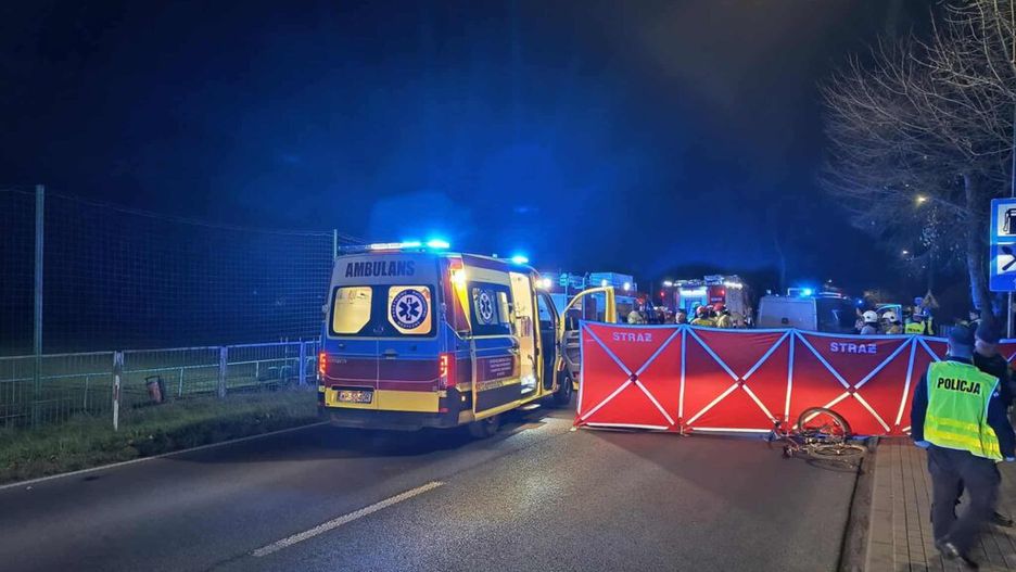 Ambulans z włączonymi sygnałami świetlnymi stoi na drodze, obok bariery zabezpieczającej miejsce zdarzenia, w nocy.