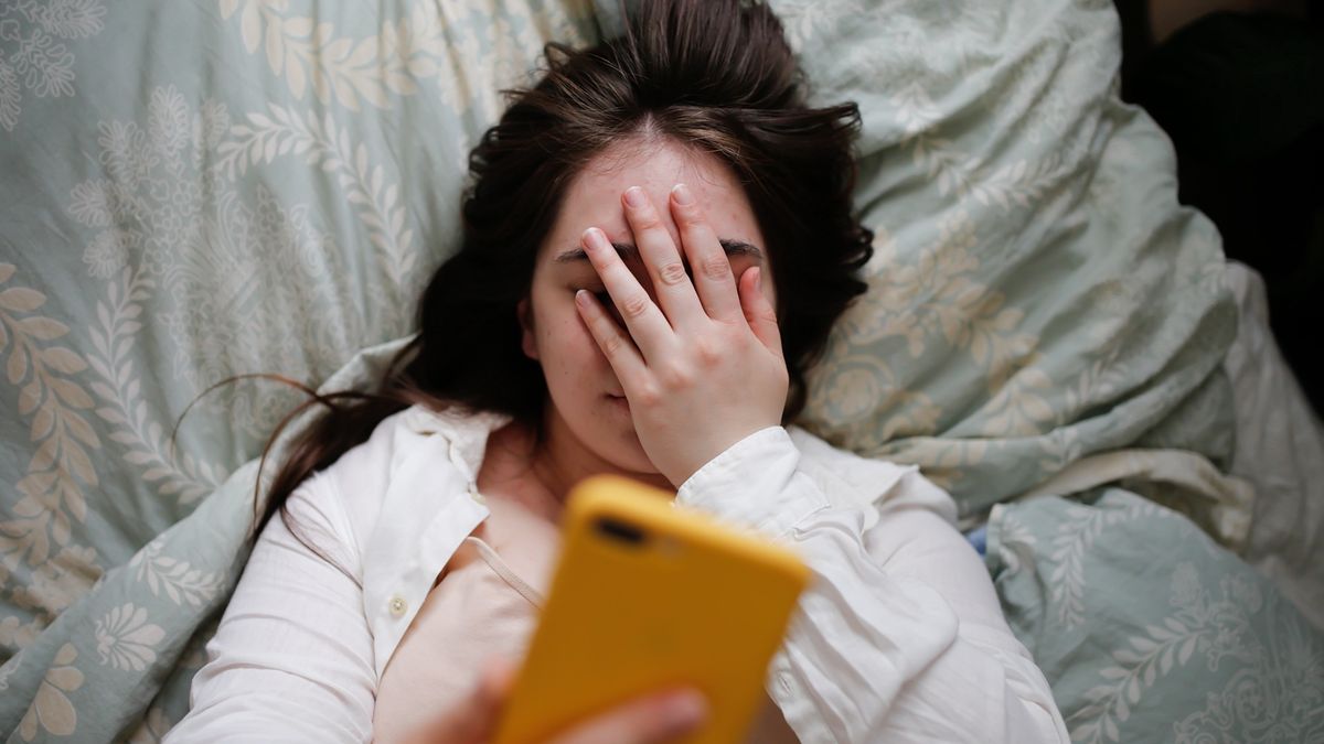 Sleepy overweight young woman with flowing hair using phone on bed, close face palm. Top viewPlus size young woman in home bedroom on bed with phone. Woman covering her eyes with her handNatalia  Lebedinskaia