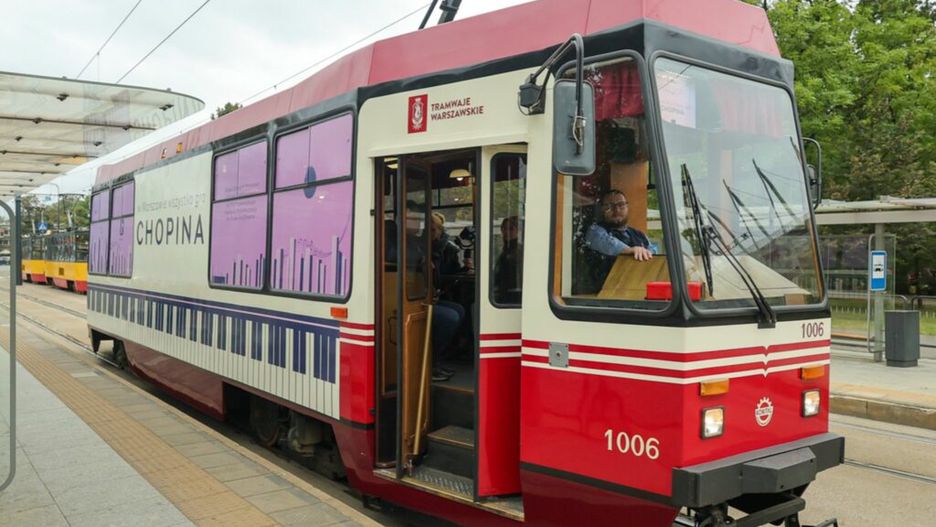 Tramwaj w barwach czerwono-białych zatrzymany na przystanku, z pasażerami wsiadającymi i wysiadającymi.