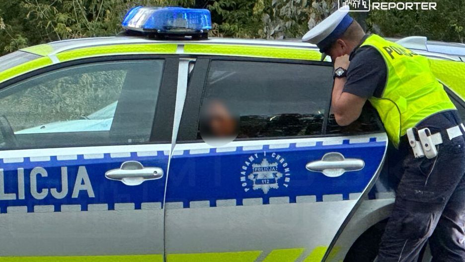 Policjant w kamizelce odblaskowej rozmawia z osobą siedzącą w radiowozie policyjnym.