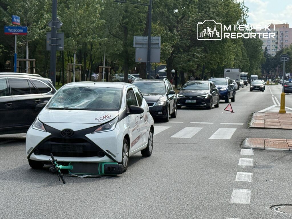 Uszkodzony biały samochód osobowy z pękniętą szybą stoi na jezdni, obok ustawiony trójkąt ostrzegawczy.