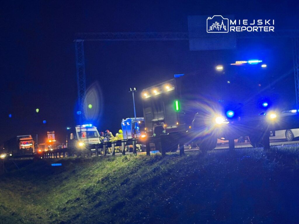 Grupa ratowników w odblaskowych kamizelkach pracuje przy wypadku drogowym, otoczona pojazdami z włączonymi sygnałami świetlnymi.