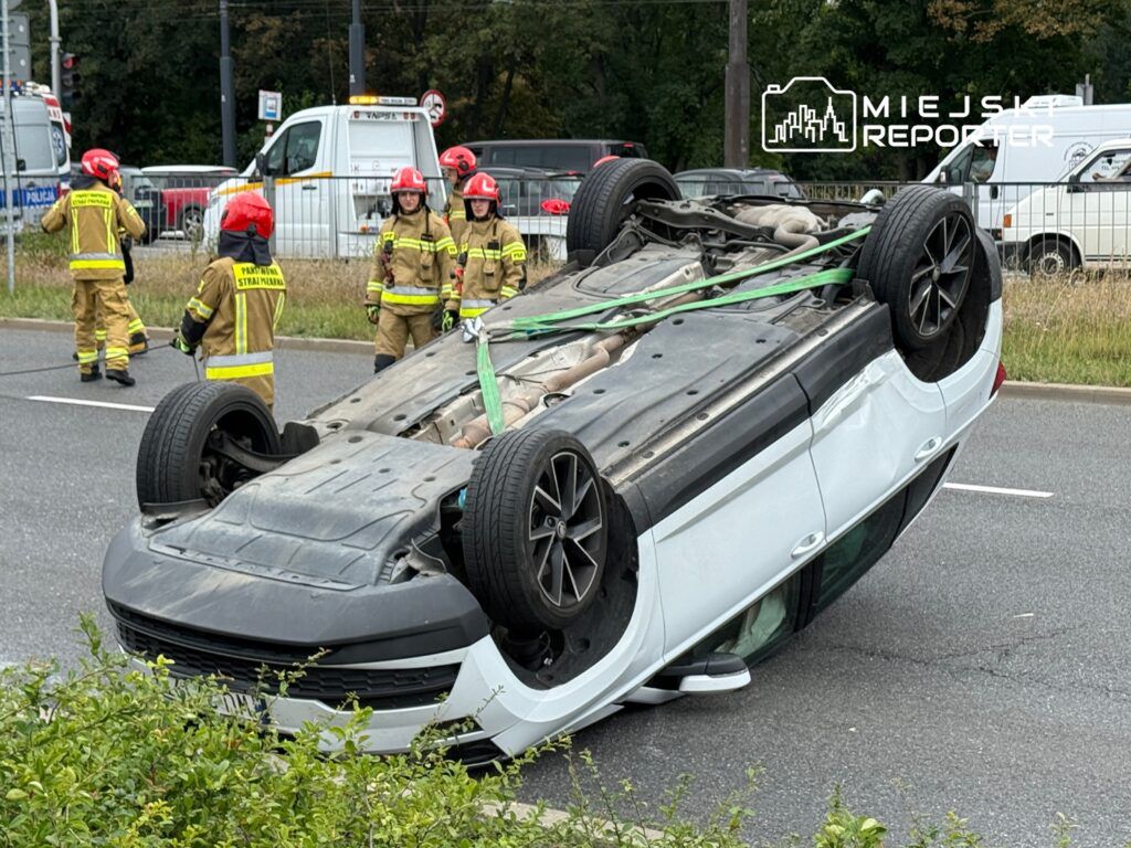 Kierowca w samochodzie osobowym leżącym na dachu, obok strażacy w mundurach pracujący przy akcji ratunkowej na drodze.