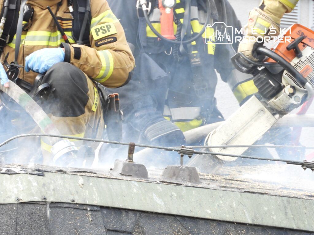 Strażacy w mundurach, w otoczeniu dymu, używają piły do cięcia dachu budynku.