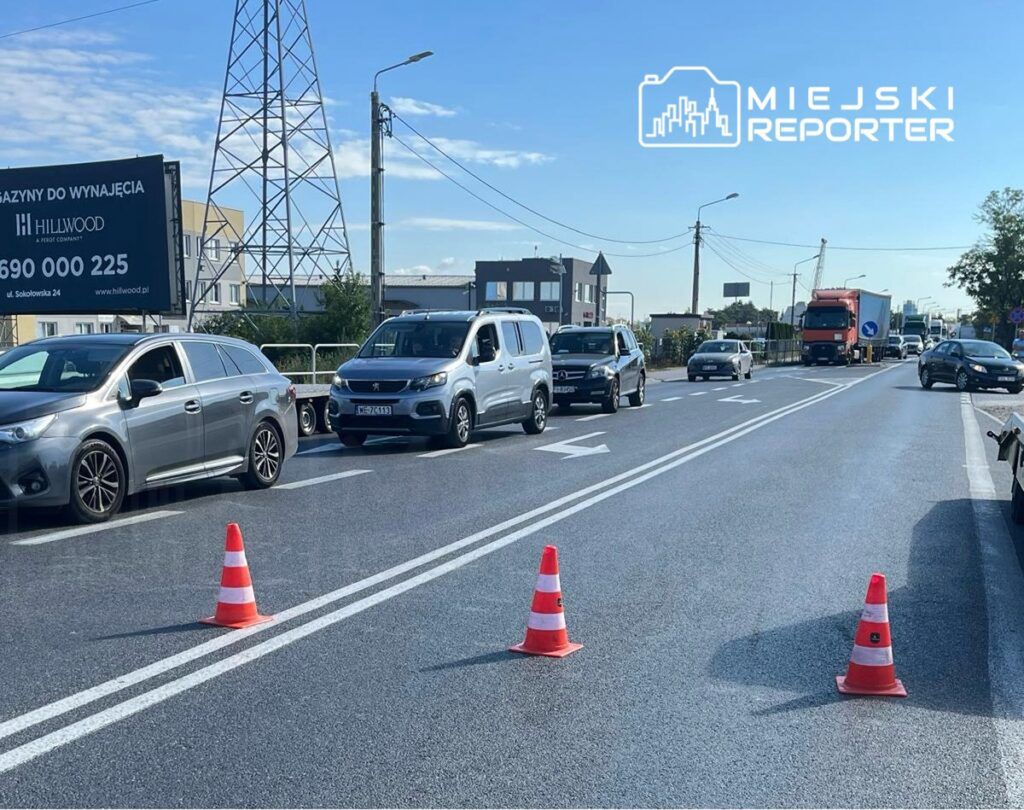 Ruch samochodowy na drodze z ustawionymi pachołkami, w tle nowoczesne budynki i reklama wynajmu magazynów.