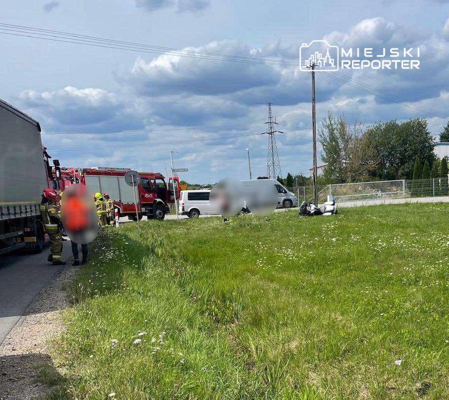 Grupa strażaków w kaskach pracuje przy drodze, obok stojącego samochodu dostawczego i wozu strażackiego.