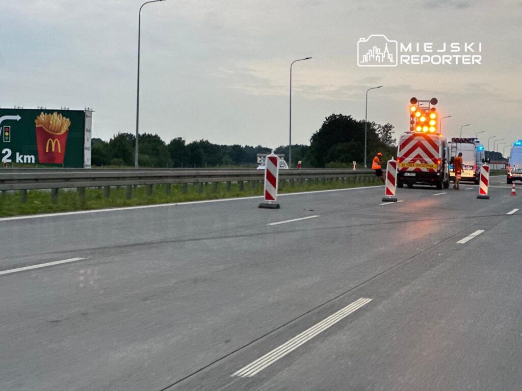 Na drodze znajdują się robotnicy w pomarańczowych kamizelkach, ustawiający znaki drogowe obok pojazdów służb drogowych.