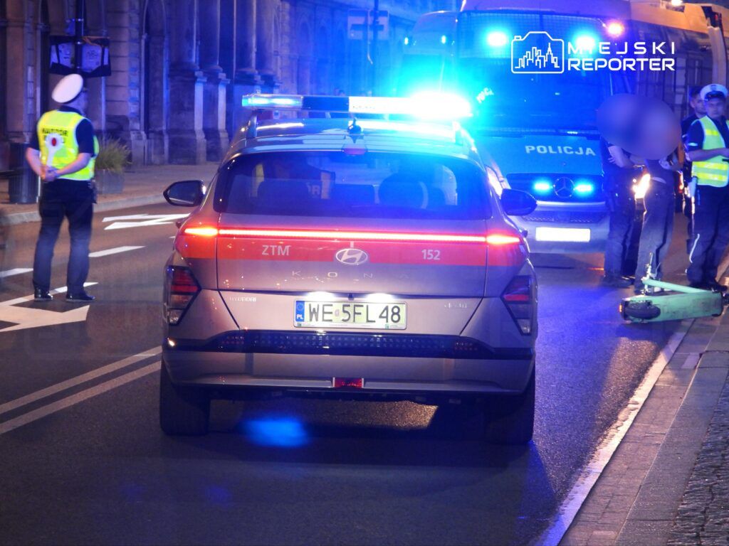 Policyjny radiowóz z włączonymi sygnałami świetlnymi stoi na ulicy, obok funkcjonariusze rozmawiają z mężczyzną.