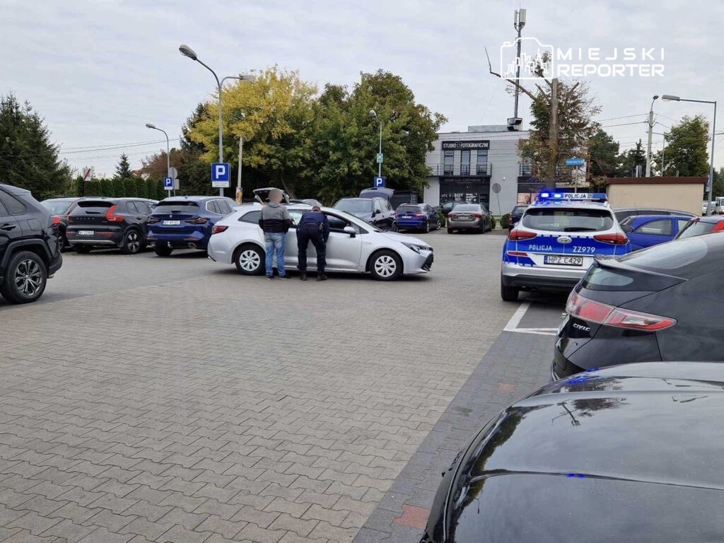 Mężczyzna w dżinsach i koszuli rozmawia z policjantem obok białego samochodu na parkingu.