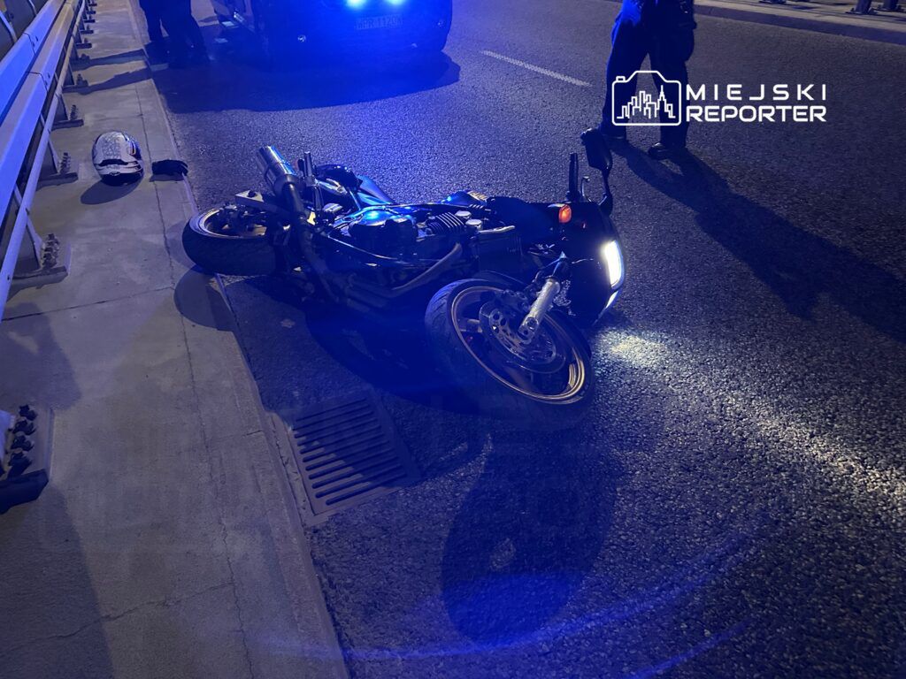 Motocykl leżący na poboczu drogi, otoczony przez policyjne radiowozy z włączonymi sygnałami świetlnymi.