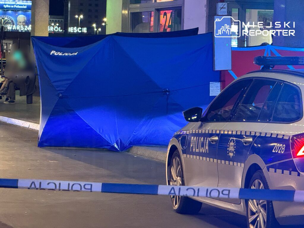 Policjant przy niebieskiej osłonie zabezpieczającej miejsce zdarzenia nocą, obok radiowozu policyjnego.