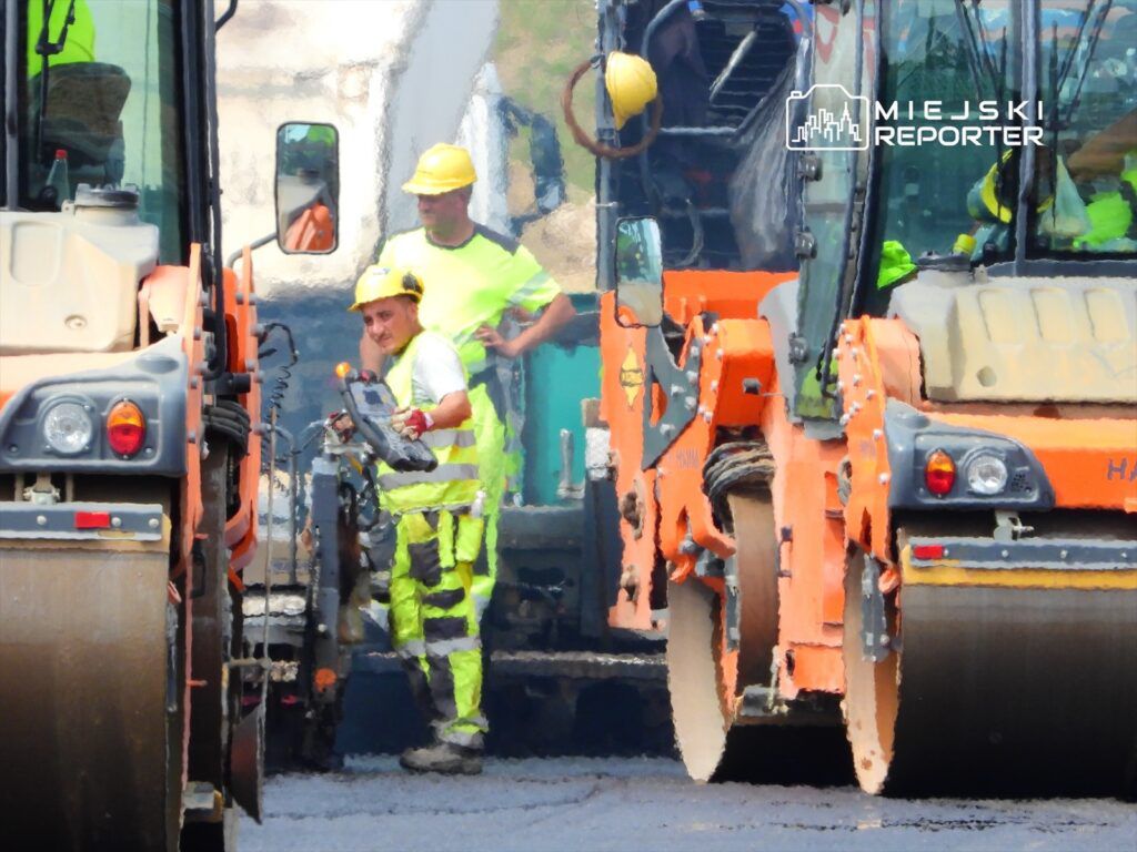 Mężczyzna w żółtej kamizelce i kasku obsługujący maszynę budowlaną na placu robót drogowych.