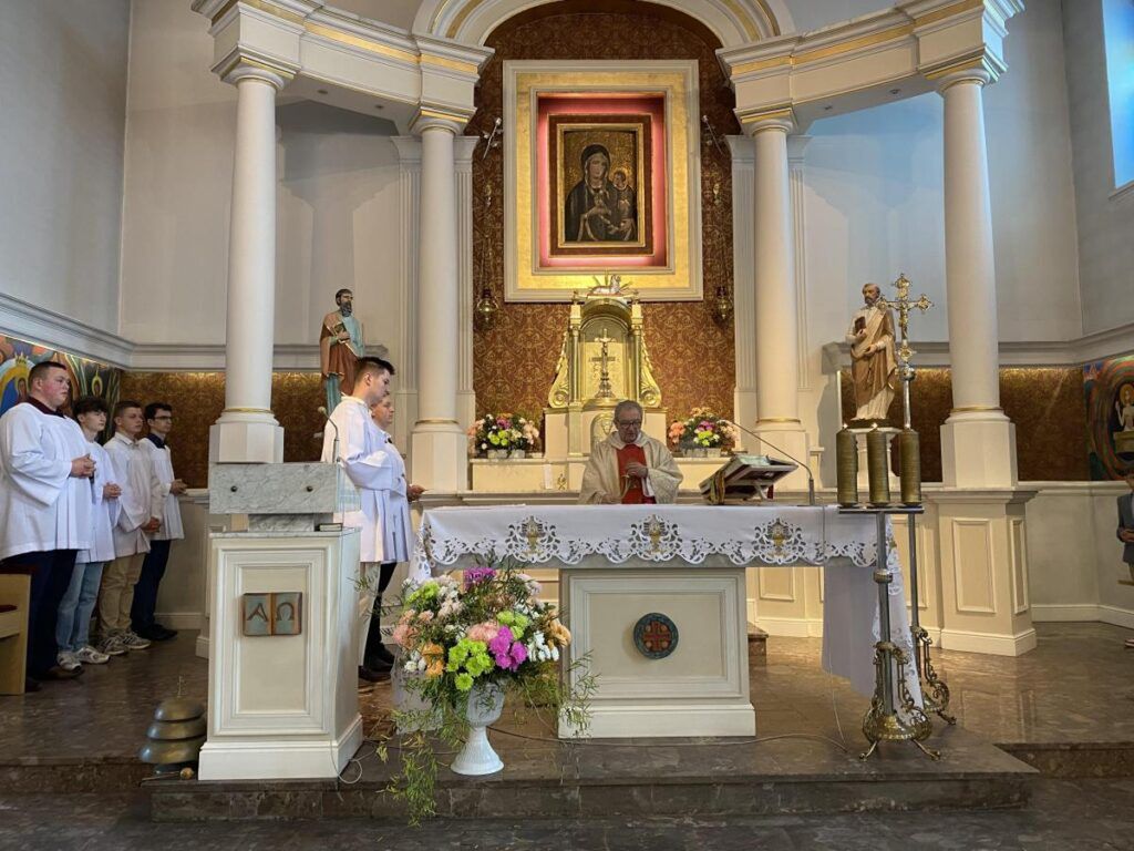 Ksiądz w szatach liturgicznych odprawia mszę przy ołtarzu w kościele, a wierni stoją w skupieniu.