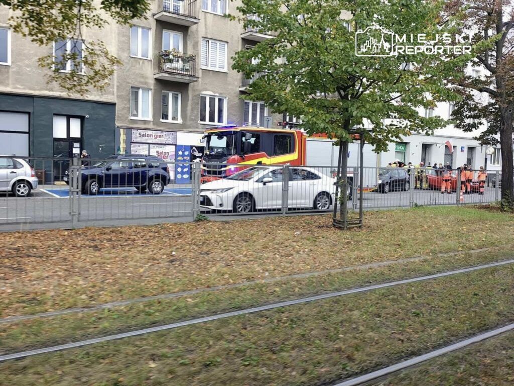 Grupa strażaków oraz pracowników w kamizelkach odblaskowych pracuje przy pojeździe przed budynkiem w miejskim otoczeniu.