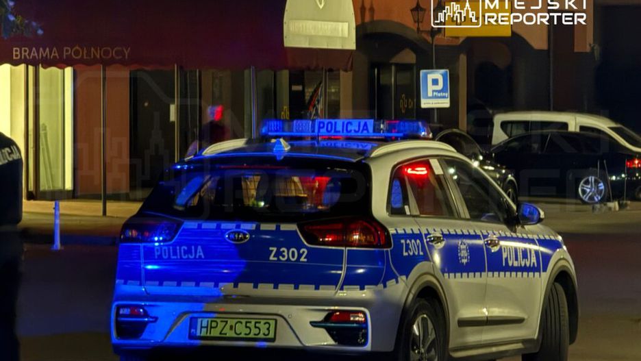 Policyjny radiowóz z niebieskimi światłami zaparkowany przed budynkiem z czerwonym daszkiem.