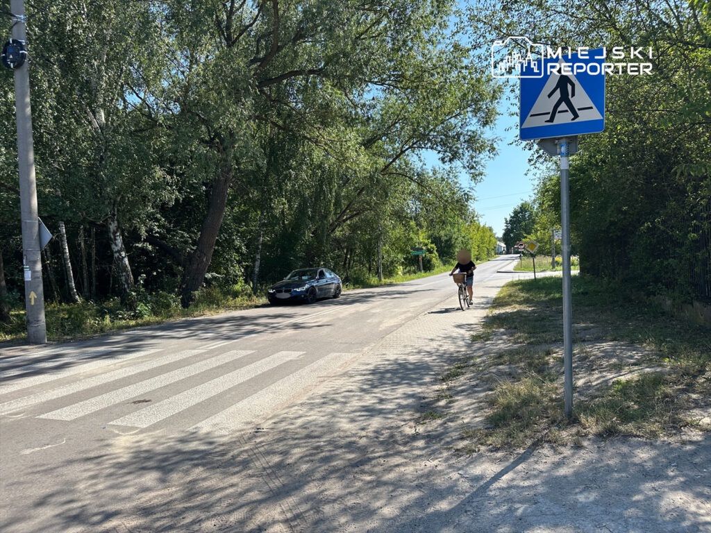 Rowerzysta jadący wzdłuż drogi obok oznakowanego przejścia dla pieszych, w otoczeniu drzew.