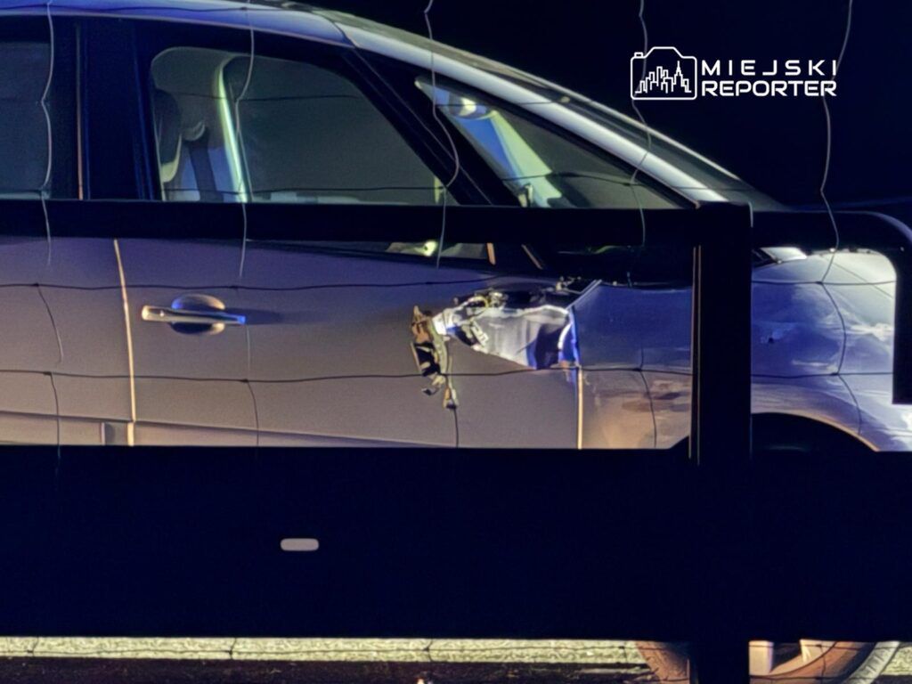 Uszkodzony samochód zaparkowany obok ogrodzenia, z widoczną wgniecioną karoserią po stronie kierowcy.