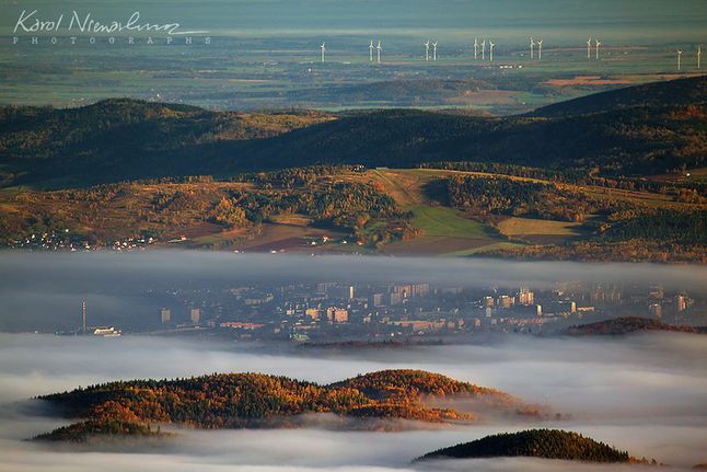 „Chyba najciekawsze ujęcie Jeleniej Góry jakie udało mi się dotychczas sfotografować! Pierwszy plan od ostatniego dzieli ponad 40 km! A samo zdjęcie zostało wykonane z głównej grani Karkonoszy - z okolicy skały Słonecznik, czyli jeszcze 10 km dalej. Ponad Wzgórzami Łomnickimi (na dole) widać wyłaniające się z porannych mgieł Kościół Łaski (zielona wieża na środku) i blokowisko "Zabobrze". Ponad mgłami pojedyncze domki Jeżowa Sudeckiego i dalej Góra Szybowcowa z charakterystycznym hangarem, w którym stoją szybowce. Ciemny wał górski na kolejnym planie to Chrośnickie Kopy, a całość zamykają równiny leżące na rubieżach Sudetów - z farmą wiatrową w rejonie Legnicy i Złotoryi" - opisuje zdjęcie autor.