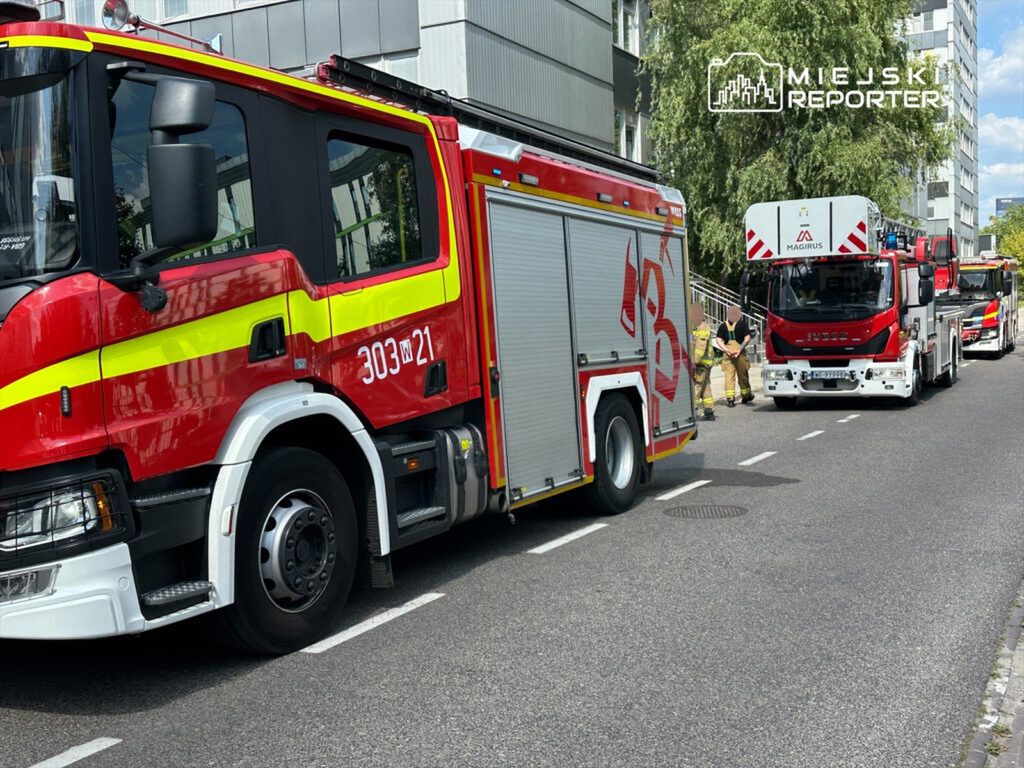 Czerwony wóz strażacki z oznaczeniem 303 w 21, zaparkowany na ulicy obok drugiego wozu, strażacy przygotowują się do akcji.
