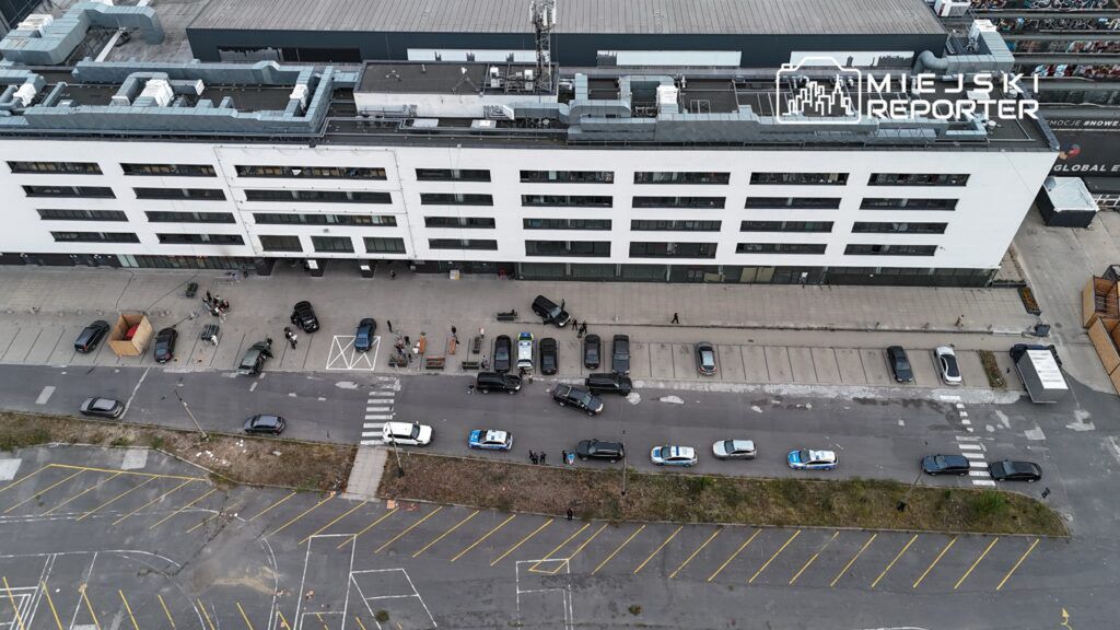 Grupa ludzi z samochodami na parkingu przed nowoczesnym biurowcem, w tle policyjny radiowóz.