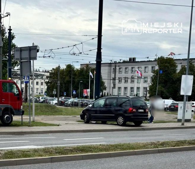 Czarny samochód zaparkowany na chodniku obok mężczyzny siedzącego na trawie w miejskim otoczeniu.