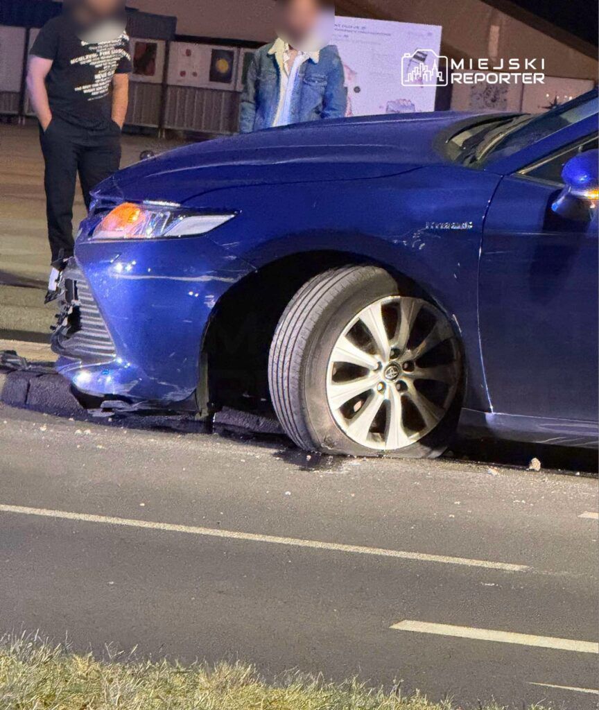Niebieski samochód osobowy z uszkodzoną oponą stoi na drodze, obok dwóch mężczyzn rozmawiających ze sobą.