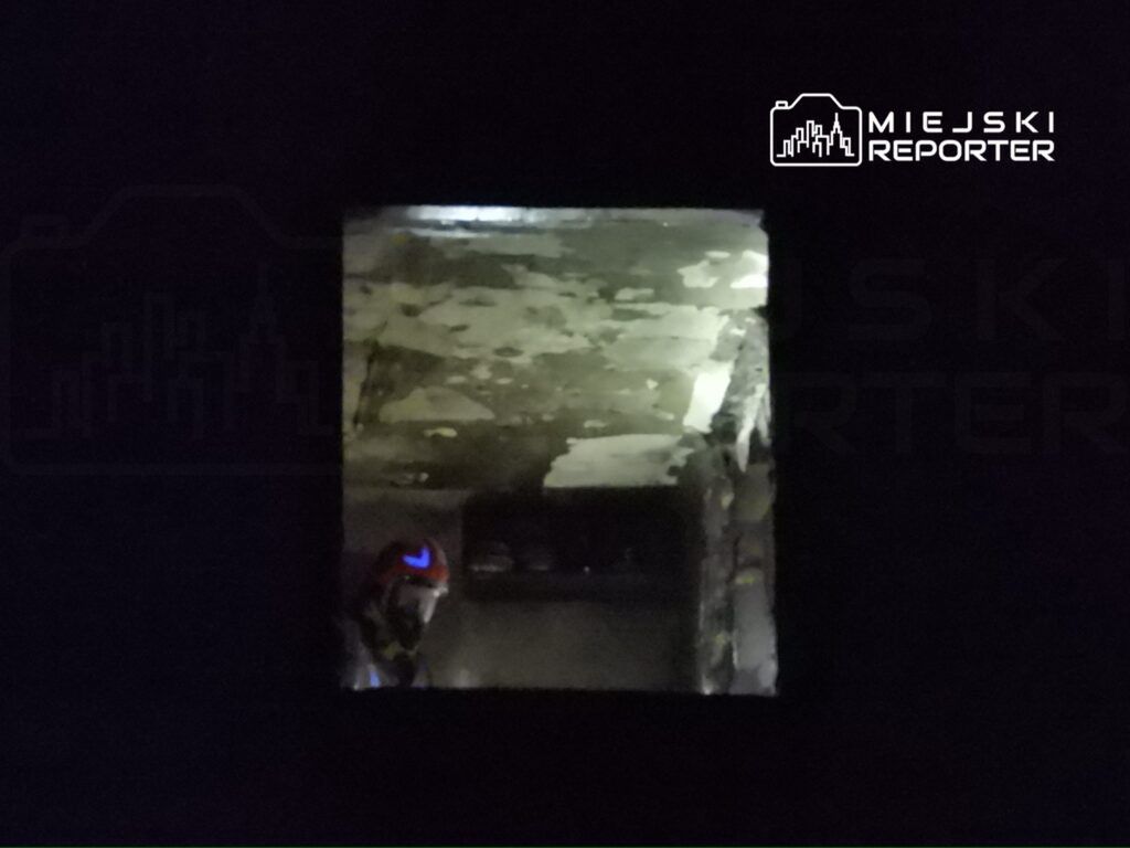 Strażak w hełmie przeszukuje ciemne, zniszczone pomieszczenie z widoczną obdrapaną ścianą.