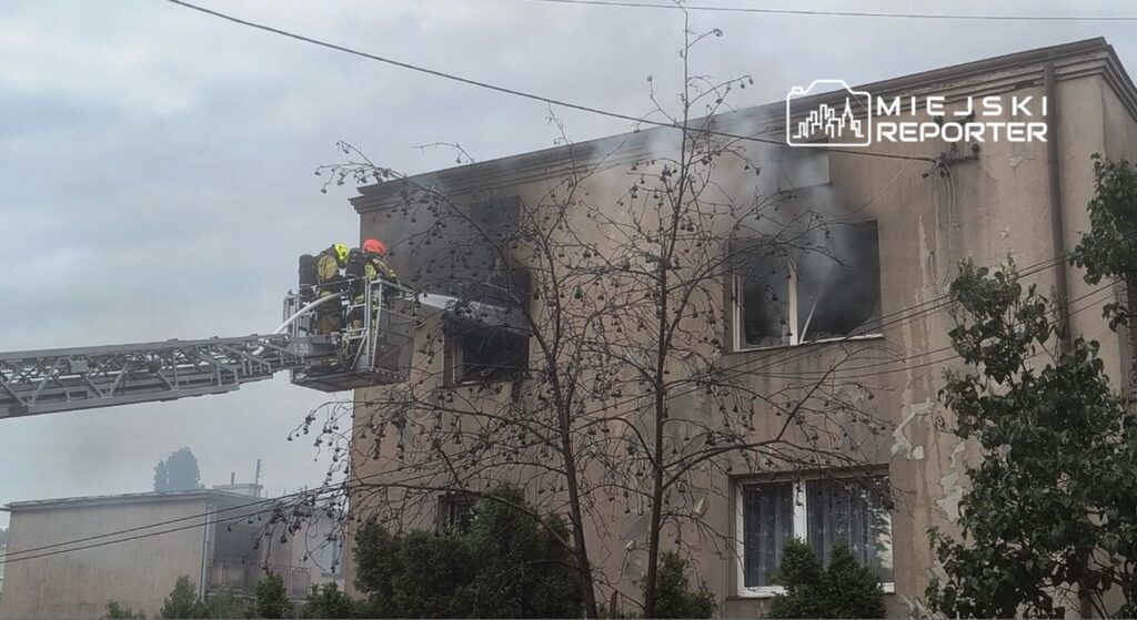 Strażacy na podnośniku gaszą pożar w oknie drugiego piętra budynku mieszkalnego. W tle widać dym i drzewa.