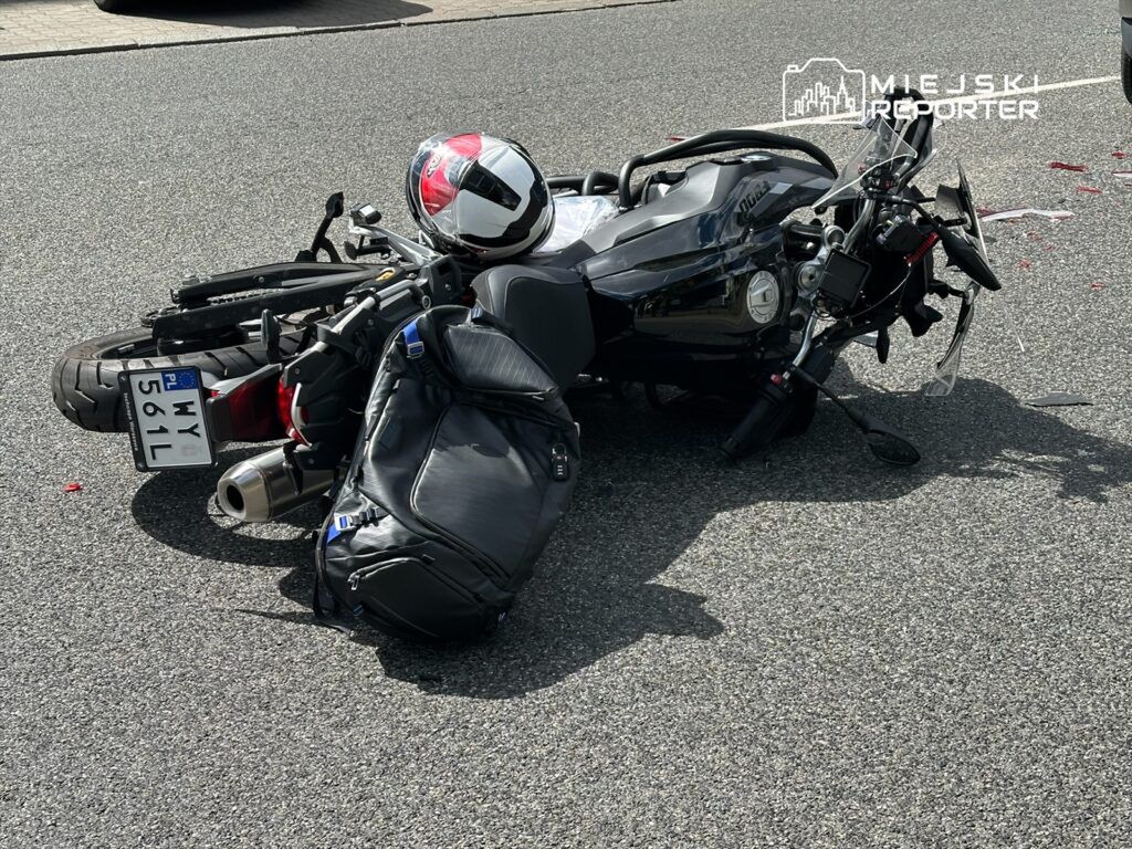Motocykl leżący na jezdni z uszkodzoną kierownicą i kaskiem obok, w otoczeniu asfaltowej drogi.