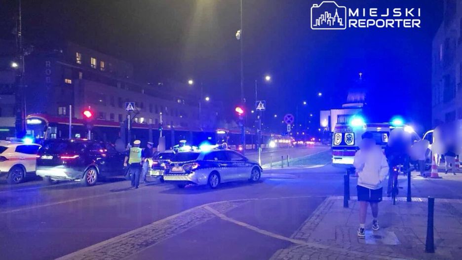 Policjanci kierują ruchem na skrzyżowaniu, obok zatrzymane pojazdy i karetka, w tle nocne miasto z oświetleniem.