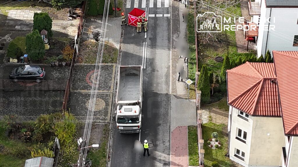 Grupa strażaków rozkłada czerwoną namiotową osłonę na ulicy, podczas gdy policjant kieruje ruchem.
