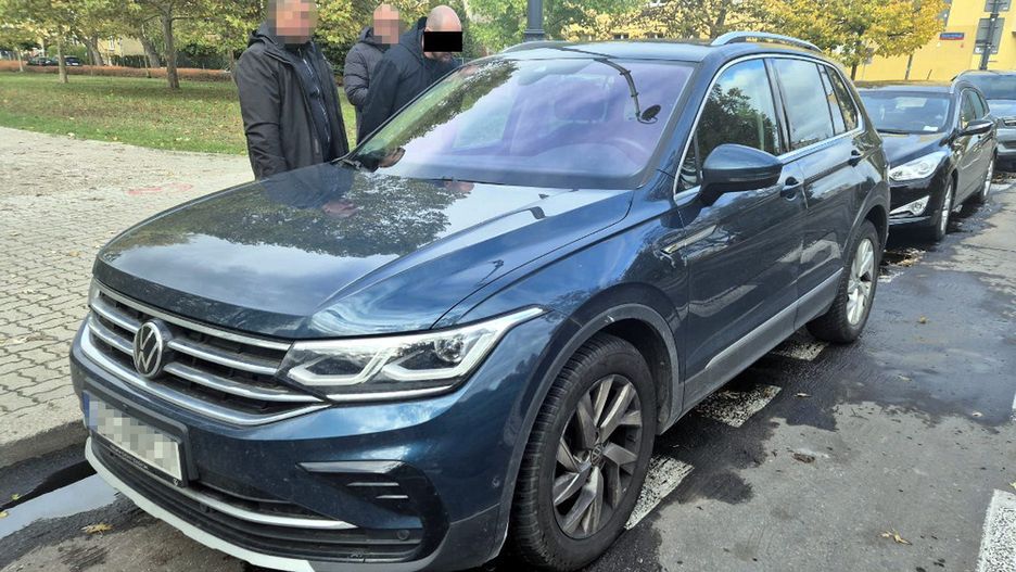 Trzech mężczyzn rozmawia obok niebieskiego samochodu marki Volkswagen zaparkowanego na ulicy, w tle drzewa.
