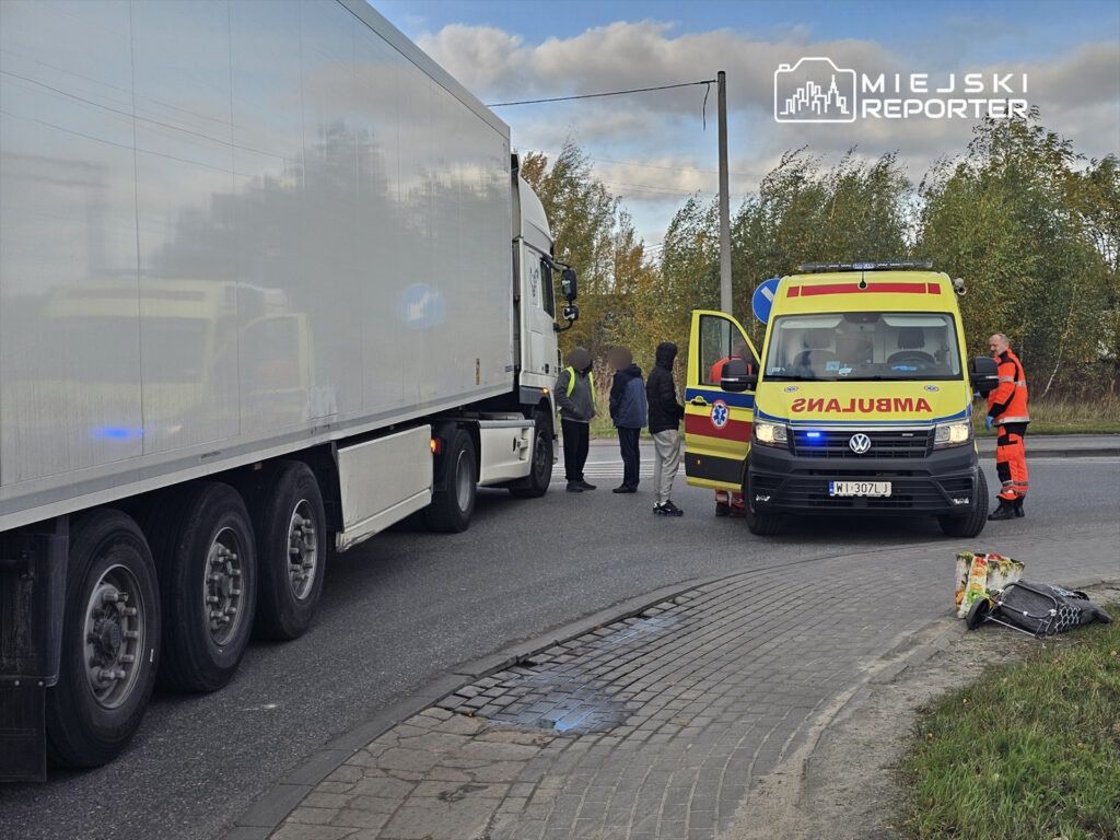 Karetka pogotowia zatrzymana przy drodze, obok stoją mężczyźni rozmawiający z ratownikami, w tle ciężarówka.
