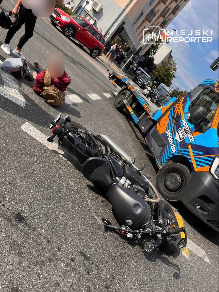 Wypadek drogowy z udziałem motocykla i osoby poszkodowanej. Motocykl leży na jezdni, a obok znajduje się ranny mężczyzna. Na miejscu zdarzenia obecni są świadkowie oraz służby ratunkowe. W tle widoczny pojazd pomocy drogowej oraz ruch samochodowy.