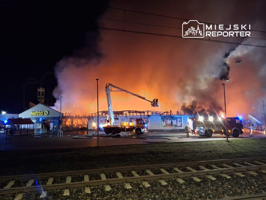 Pożar w sklepie Netto, gdzie strażacy walczą z ogniem przy użyciu podnośnika i wozów strażackich w nocy.
