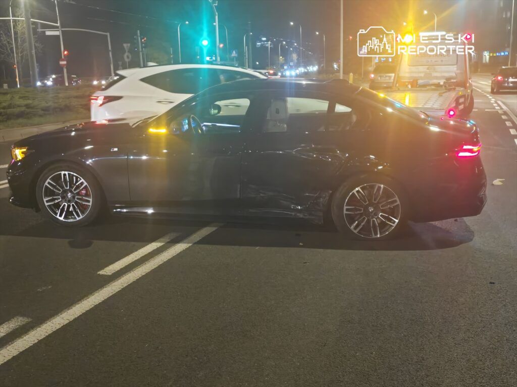 Czarny sedan z uszkodzeniem boku stoi na drodze, obok przejeżdżają inne pojazdy w nocy.