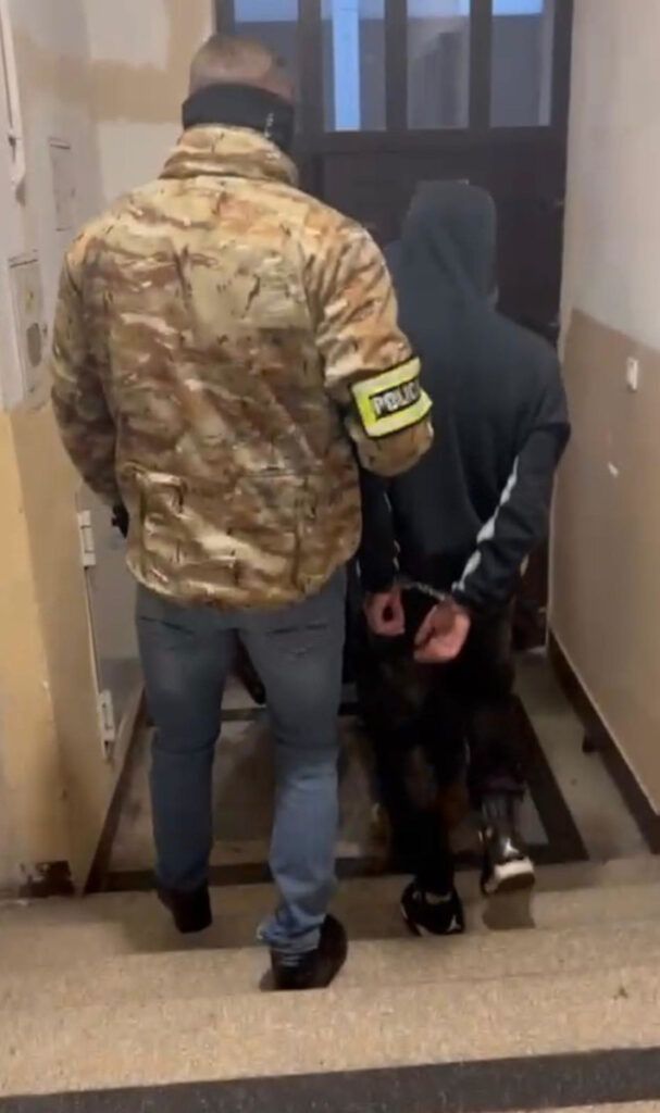 Funkcjonariusz w kamizelce policyjnej eskortuje zatrzymanego mężczyznę schodami w budynku.