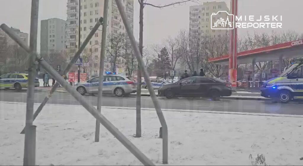 Policjanci prowadzący dochodzenie obok zaparkowanych radiowozów na zaśnieżonej ulicy w mieście.