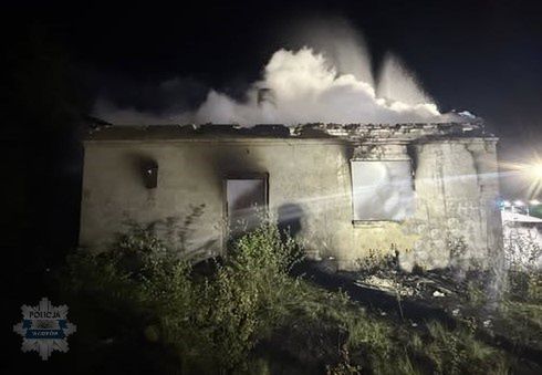 Dom zniszczony przez pożar, z którego wydobywa się dym, otoczony roślinnością w nocy.