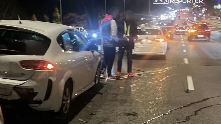 Dwaj mężczyźni rozmawiają obok uszkodzonego białego samochodu na ruchliwej drodze nocą.