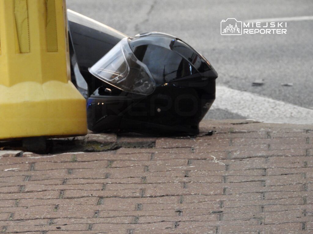Czarny kask motocyklowy leży na chodniku obok żółtej bariery, w tle widoczna ulica.