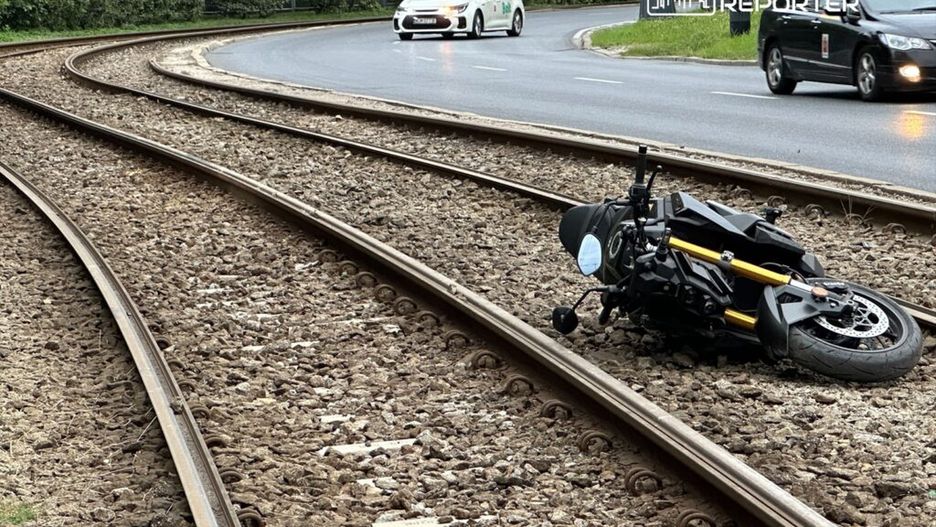 Motocykl leży na torach tramwajowych, obok ruchliwej ulicy, w otoczeniu zieleni i samochodów.