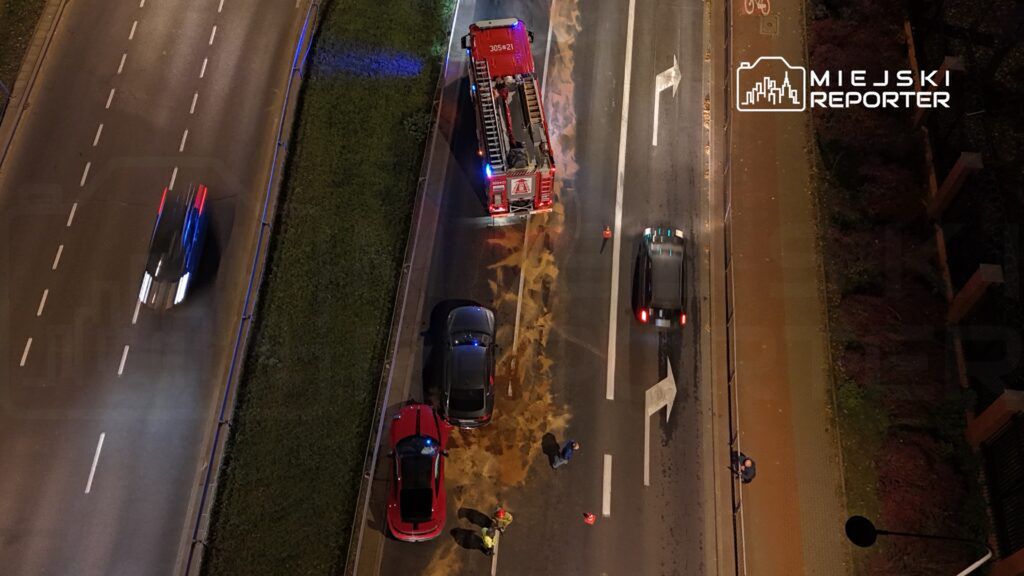 Na jezdni znajdują się dwa uszkodzone samochody, a obok nich straż pożarna usuwająca plamę substancji.